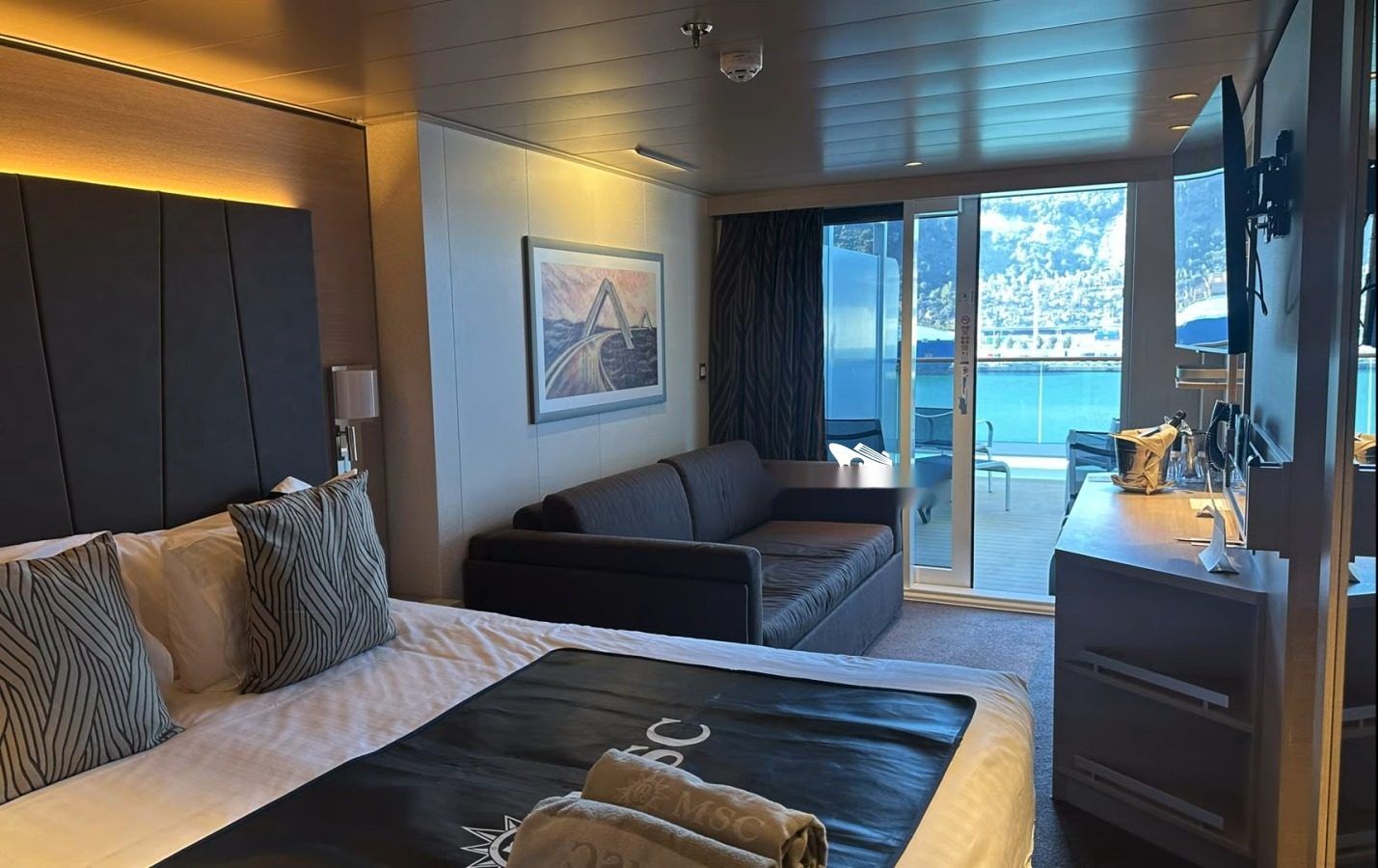 Каюта с балконом (Balcony) на MSC World Europa — кровать, диван и выход на балкон с видом на порт
